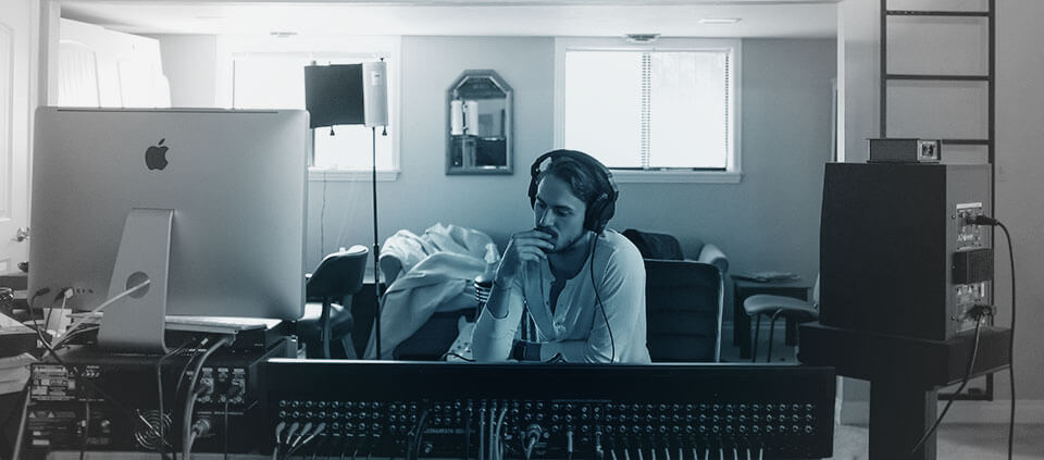 man sitting in his room with a modern music recording setup, with a contemplative look on his face 